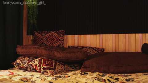 Media: Video of a cozy, dimly-lit bedroom with a bed covered in rich, patterned bedding featuring red and gold designs. The walls are wooden paneling, and the window is covered with dark blinds. The room exudes warmth and comfort.