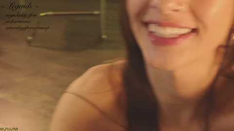 Video of a smiling woman with fair skin and dark hair, wearing a strapless top, taken indoors. Background shows a blurred, dimly lit room with metallic objects.