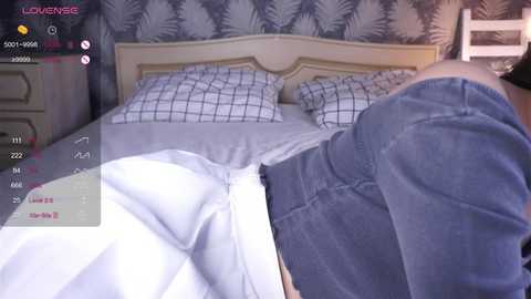 A video captures a woman's exposed buttocks in blue jeans, lying on a bed with a gray duvet and white pillowcases. The background features a wooden headboard and a wallpaper with a leafy pattern.