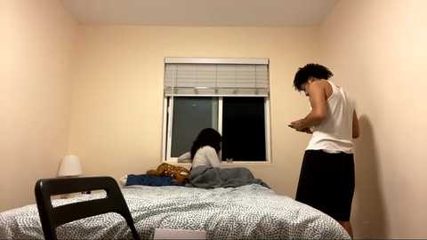 Media: Video of a small, dimly lit bedroom with beige walls. A young woman with long hair, wearing a white tank top, leans against a window. Another woman with short hair, dressed in a grey shirt, lies on a bed with a patterned blanket.