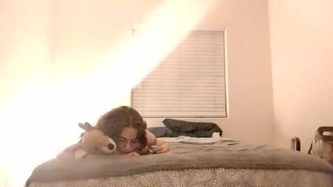 Media: Video of a young woman with shoulder-length brown hair lying on a bed, cuddling a stuffed dog, in a dimly lit room with a window covered by white blinds.