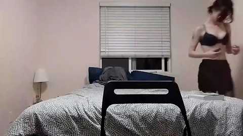 Media: Video of a woman in a black bra and skirt adjusting her bra in a small, dimly lit bedroom with a bed, patterned bedspread, closed window blinds, and a lamp.