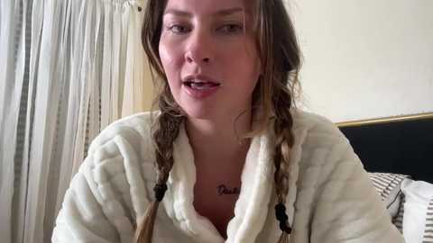 Video of a young Caucasian woman with light skin, brown hair in two braids, wearing a white bathrobe, with a tattoo on her chest, speaking in a bedroom with beige curtains and a black and white striped pillow.