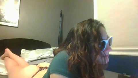 Media: A video of a woman with wavy brown hair and large sunglasses lying on a bed in a dimly lit room with gray walls and a window.