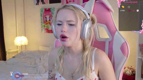 Media: Video of a young blonde woman with fair skin, wearing a pink and white floral-patterned bra, sitting in a pink gaming chair, looking tired, with a white lamp and posters in the background.