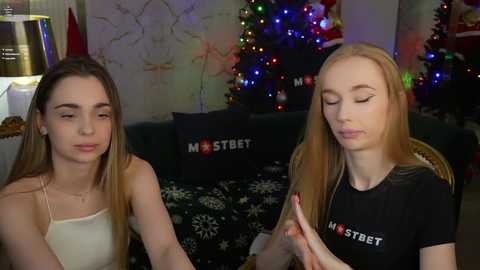 Media: Video of two young women with long hair, one in a white top, the other in a black shirt, seated indoors, with a Christmas tree and decorations in the background.