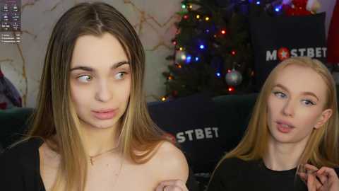 Media: Video of two young women: one with long brown hair, fair skin, wearing a black top, and the other with blonde hair, fair skin, and a black top, both in a cozy indoor setting with a decorated Christmas tree in the background.