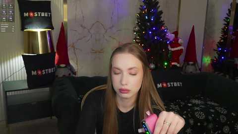 Media: Video of a young woman with long brown hair, wearing a black hoodie, holding a pink M&S toy in a cozy living room with a Christmas tree and decorations.