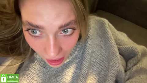 Media: Video of a young woman with fair skin, green eyes, and long blonde hair, wearing a grey knitted sweater, looking shocked or surprised. Background is blurred.