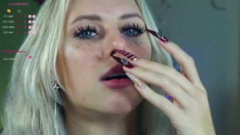 Video of a fair-skinned, blonde woman with blue eyes, long eyelashes, and glossy red lipstick, applying dark red nail polish on her nails. The background is blurred with green and purple hues.