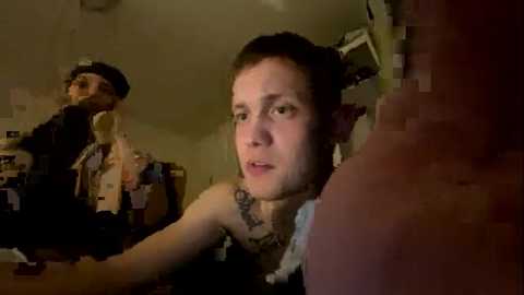 Media: Video of a pale, tattooed young man with long, dark hair in a messy bun, wearing a black tank top, sitting on a cluttered couch in a dimly lit room.