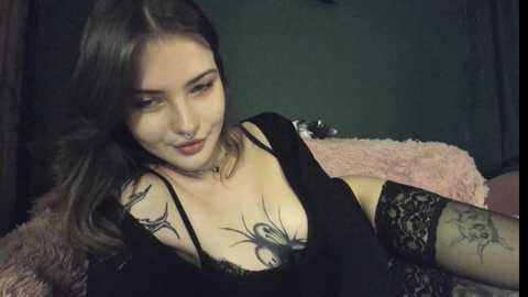 Media: Video of a young woman with long dark hair, fair skin, and black lace lingerie, lying on a pink fluffy bed. She has tattoos on her chest and arms.