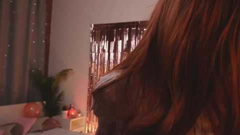 Video of a woman with long, reddish-brown hair, leaning on a bed with a white duvet, against a backdrop of a shiny curtain and a green potted plant.