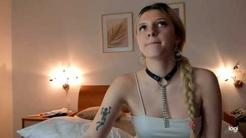 Media: Video of a fair-skinned woman with long blonde hair, wearing a black leather choker and a white spaghetti-strap top, sitting on a bed with a tattoo on her left arm, in a dimly-lit, cozy bedroom with wall sconces and framed artwork.