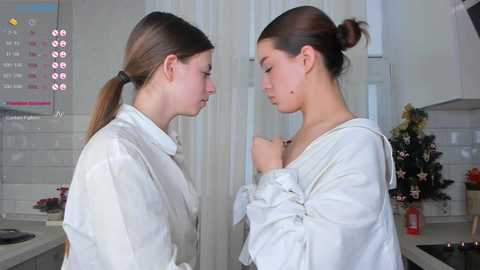 Media: Video of two young women with light skin, brown hair tied back, wearing white robes, facing each other in a modern kitchen.