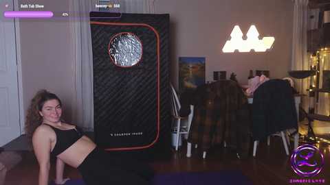 Video of a pregnant woman with curly hair in a black sports bra, leaning against a black exercise ball. Background features a gaming setup, a white door, and a plush rug.