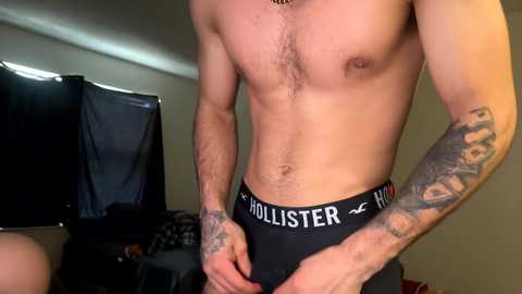 Media: Video of a shirtless man with tattoos wearing black Hollister briefs, standing indoors near a TV and dark cloth.