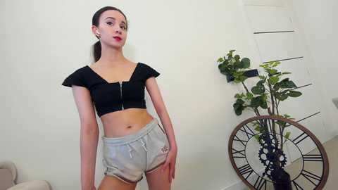 Media: Video of a slim, fair-skinned woman with brown hair, wearing a black cropped top and grey shorts, standing in a modern, minimalist room with a white wall, a potted plant, and a vintage-style clock.