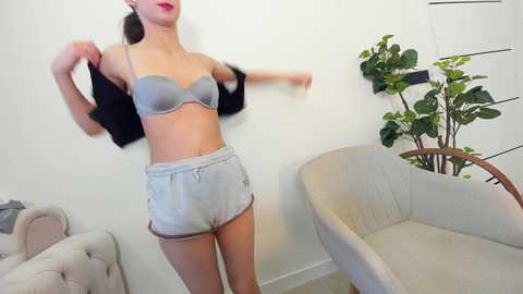 Media: Video of a slim, fair-skinned woman with red lipstick, wearing a light gray bra and shorts, lifting a black jacket, standing in a modern, minimalist room with a beige armchair, potted plant, and white wall.