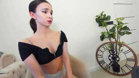 Media: Video of a young Asian woman with fair skin and long black hair, wearing a black off-shoulder bra and light gray skirt, sitting on a beige couch in a minimalist room with a large clock and green plants.