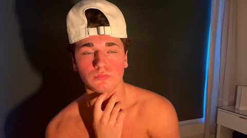 Video of a shirtless young man with short brown hair, wearing a backwards white baseball cap, looking contemplative with his hand on his chin. Background shows dimly lit room with dark green walls and a white window.