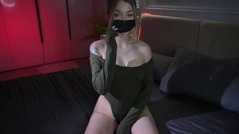 Video of a slender, fair-skinned woman with long brown hair, wearing a black face mask and olive-green off-shoulder bodysuit, sitting on a dark bed with gray pillows, in a dimly lit, modern bedroom.