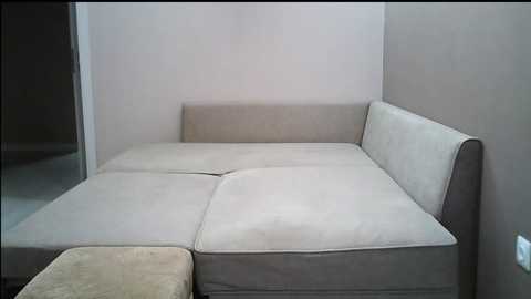Media: Video of a minimalist living room corner with a beige sectional sofa against a light gray wall, featuring clean, straight lines and simple, modern design.