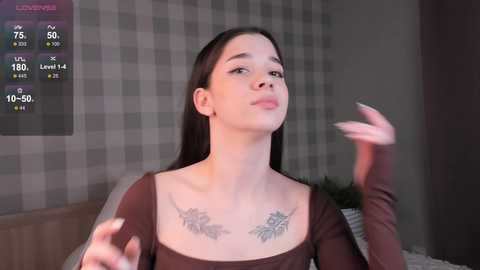 Video of a young woman with pale skin and long dark hair, wearing a brown top, with two floral tattoos on her chest. She gestures with her hands. Background includes a grey and white checkered wall.