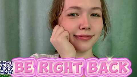 Video of a young woman with light skin and brown hair, smiling with one hand on her cheek, set against a greenish-gray background. Text reads \"BE RIGHT BACK\" in bold, pink letters.