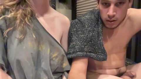 Media: Video of a young man with a shocked expression, shirtless, leaning against a woman in a see-through grey top. They are indoors, with a textured grey towel draped over the man's shoulder.