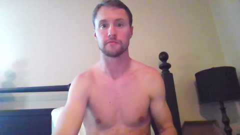 Media: A video of a shirtless, light-skinned man with short brown hair, wearing a neutral expression, standing in a bedroom with a dark wooden headboard, beige walls, and a black lampshade.