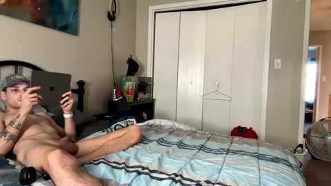 Video of a naked young man with a slender build, light skin, and tattoos, lying on a bed with a blue and gray checkered blanket, holding a smartphone, in a cluttered bedroom with a white wardrobe and a red fan.