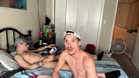 Media: Video of two shirtless men in a messy bedroom; one sits on a bed, the other stands in front, both wearing caps. Background includes a bed, white doors, and a fan.