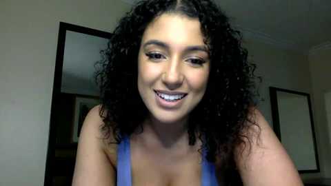 Media: Video of a smiling, curly-haired woman with light skin tone, wearing a blue tank top. She is indoors, with a neutral-colored background featuring a mirror and a window.