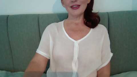 Media: Video of a middle-aged woman with fair skin and auburn hair, smiling, wearing a sheer white blouse, sitting on a green couch against a light blue wall.