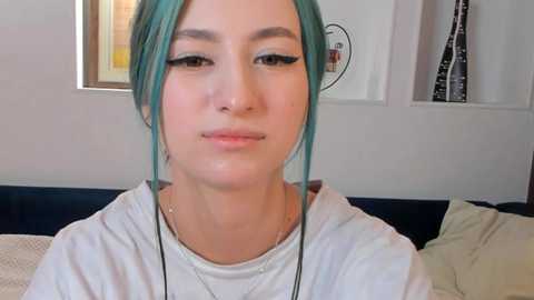 Media: Video of a young woman with teal hair, wearing a white T-shirt, with a neutral expression, and headphones around her neck, in a modern, minimalistic room with a black couch and white walls.