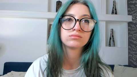 Media: Video of a young woman with light skin and long, turquoise hair, wearing large black glasses and a white shirt, seated in a modern living room with white shelves and dark furniture.