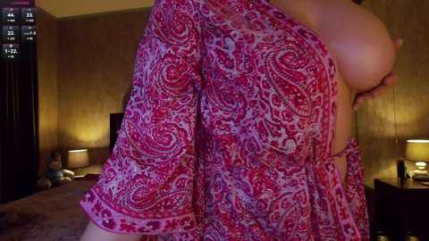 Video of a woman with fair skin, large breasts, and a red paisley-patterned robe, partially open, in a dimly lit bedroom with warm lighting and patterned wallpaper.