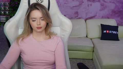 Media: Video of a young Caucasian woman with light skin and blonde hair, wearing a pink top, sitting on a white gaming chair in a purple-painted room with a beige sectional couch and a \"Masturbate\" pillow.