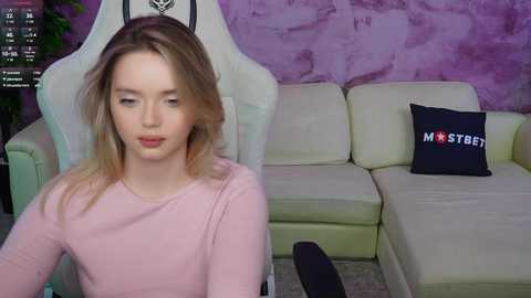 Video of a young, fair-skinned woman with shoulder-length blonde hair in a pink long-sleeved top, seated on a white gaming chair. Background features a light beige sectional couch with a black pillow reading \"MISTERB\" against a purple wall.