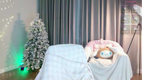 A video of a cozy room with a white blanket-covered chair, a pink and white teddy bear, a snow-covered Christmas tree, and a large light fixture.