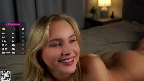 Video of a smiling, nude blonde woman with long hair lying on a bed in a dimly lit room, with a digital screen displaying a \"Live Newscast\" in the background.