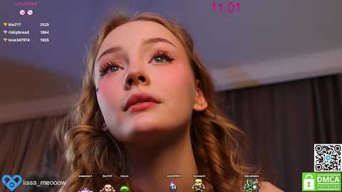 Video of a young, fair-skinned woman with wavy, light brown hair, wearing makeup. She has pink blush on her cheeks and is looking directly at the camera. Background shows a dark curtain.