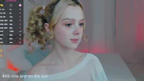 Media: A video of a young woman with blonde curly hair, wearing a sheer white top, captured in a dimly lit room with soft pink and blue lighting.