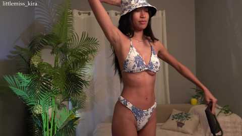 Media: Video of a slender woman with long dark hair, wearing a floral bikini and a bucket hat, posing indoors beside a potted plant.