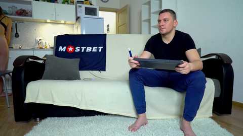 Media: Video of a clean-cut, barefoot white man in a black T-shirt and blue jeans, sitting on a beige couch, holding a tablet, in a modern, minimalist kitchen with white cabinets, a microwave, and a white refrigerator.