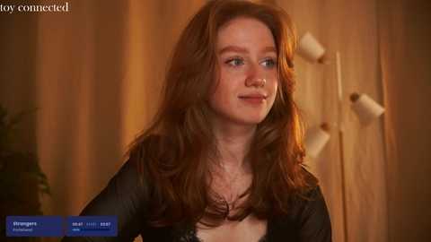 Media: Video of a fair-skinned, red-haired woman with long, wavy hair in a black top, smiling softly. Background features wooden door handles and a warm, cozy indoor setting.