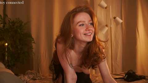 Media: Video of a smiling redhead woman with long hair, wearing a black strapless dress, in a warmly lit room with beige curtains and a green plant.