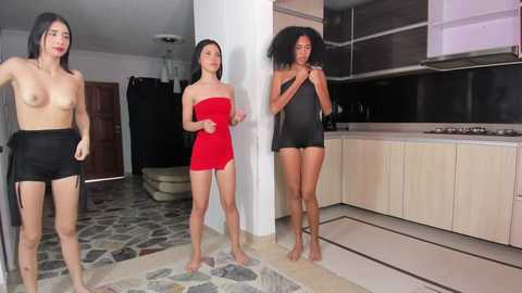 Media: Video of three topless women in a modern kitchen; two wear red and black dresses, the third is bare-chested, all with varying skin tones.