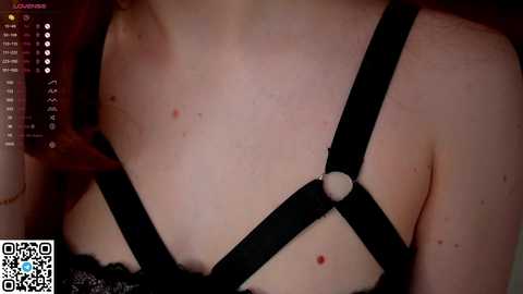 Media: A close-up video of a fair-skinned woman's neck and chest, adorned with a black leather harness. The harness features a circular ring. The background is dimly lit, with a QR code and text at the bottom left corner.